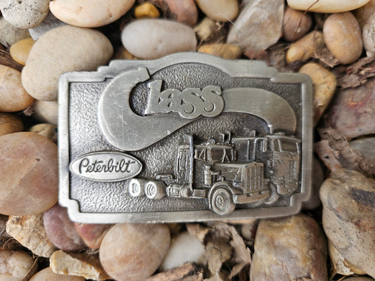 Belt buckle with the word Class, two semi trucks and a Peterbilt logo, on pebbles