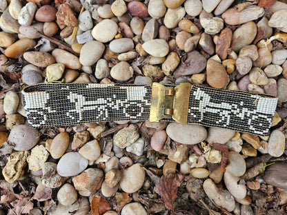 Black and white beaded stretch belt with a horse and carriage scene and a brass hinge closure, laid flat on pebbles