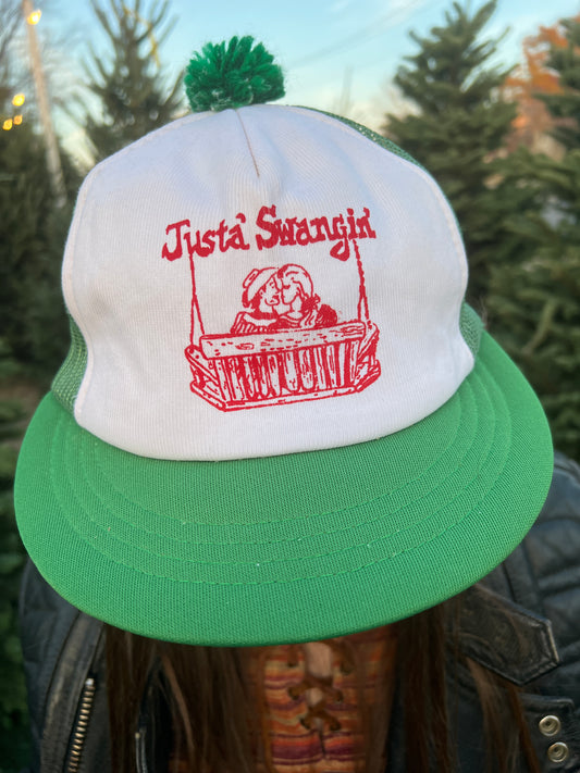 Green trucker hat with a pom pom and a porch swing couple graphic captioned 'Just a Swangin', worn by a model