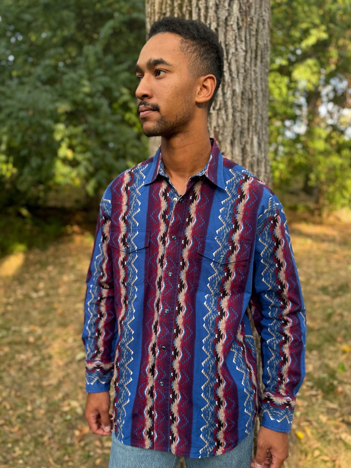 Person in a blue and burgundy patterned western shirt, standing by a tree