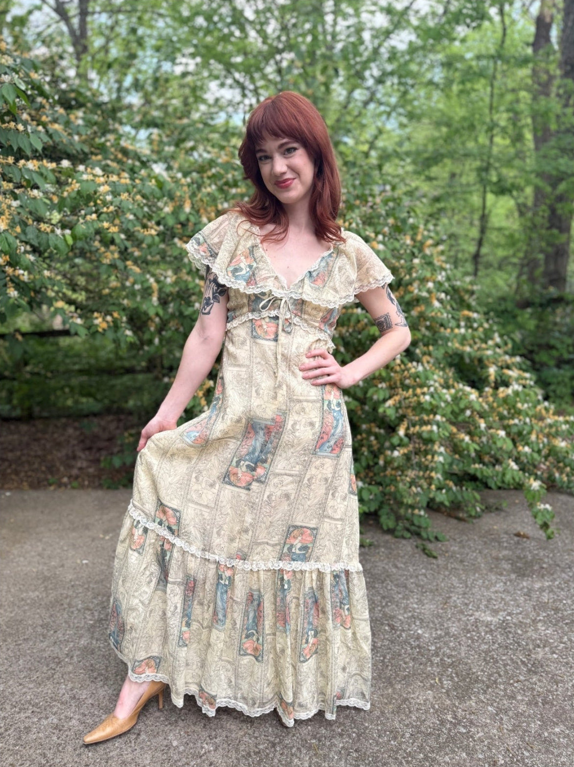 Person in a cream printed maxi dress with lace trim and a ruffled neckline, standing outdoors by green shrubs