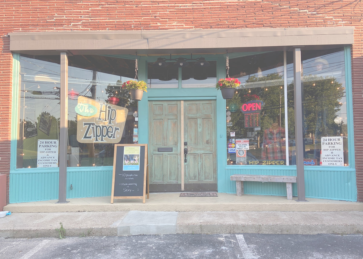 Storefront of The Hip Zipper with red brick above teal trim, green double doors and a hanging Hip Zipper sign