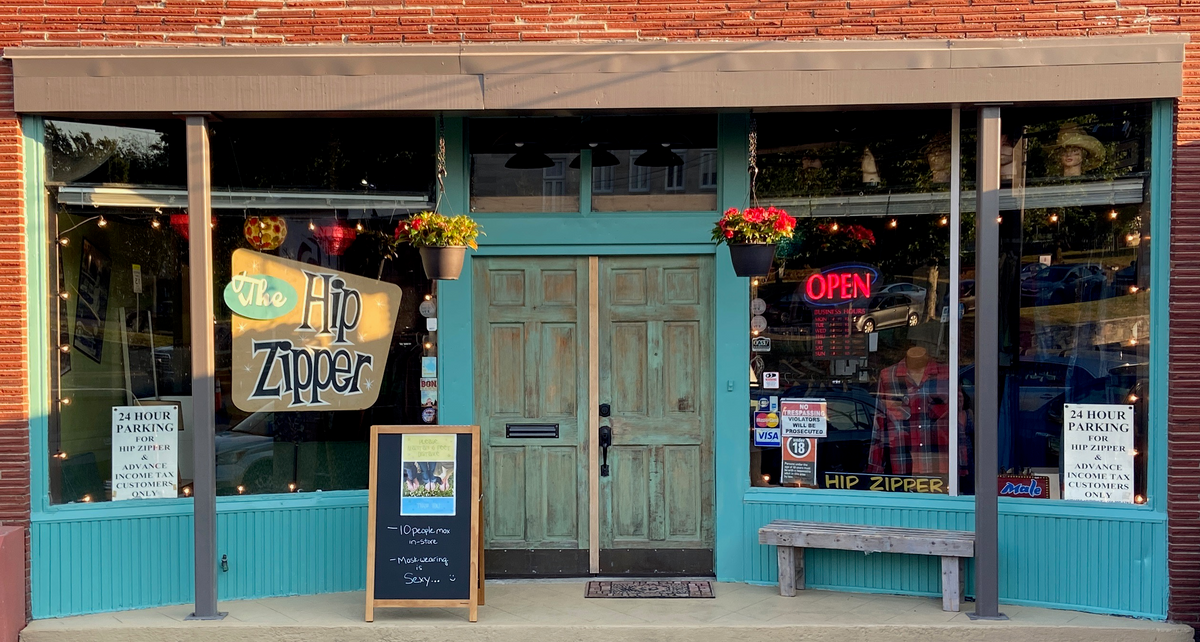Storefront of The Hip Zipper with teal trim, green double doors and a hanging Hip Zipper sign