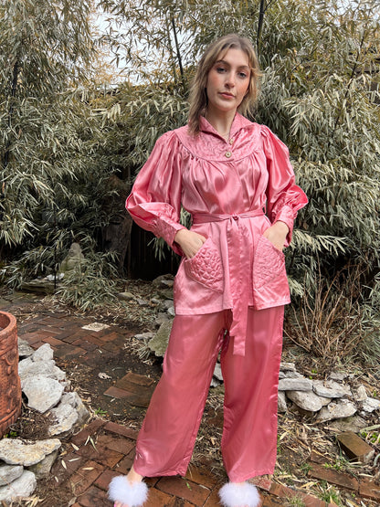 Pink satin pajama set with a belted top, bloused sleeves, quilted pockets and wide-leg pants, worn by a model