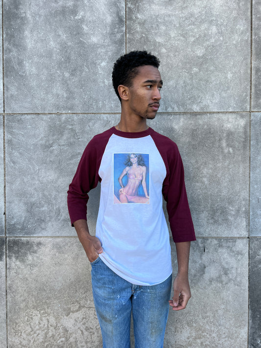 White baseball tee with three-quarter sleeves and a photo transfer of Cheryl Tiegs in a bikini, worn by a model