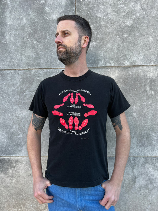 Black t-shirt with cha-cha-cha dance step instructions on the front, worn by a model with jeans