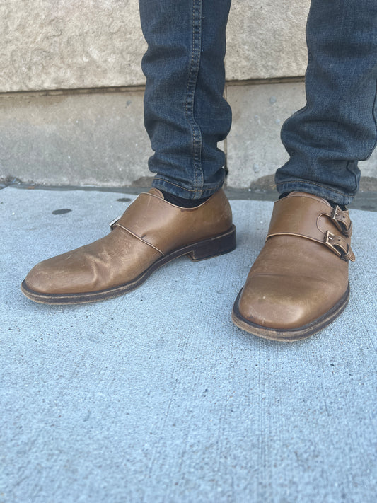Pair of brown leather dress shoes with two side buckles, worn with jeans on a sidewalk